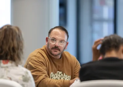 A participant in a brown sweatshirt listens to others around the Lived Experience Committee meeting table.