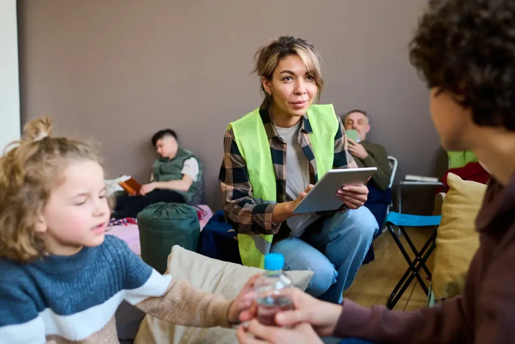 A volunteer in a high-visibility vest sits with a tablet and talks with people resting on bedding in a shelter room, while in the foreground a child is handed a bottle of water.