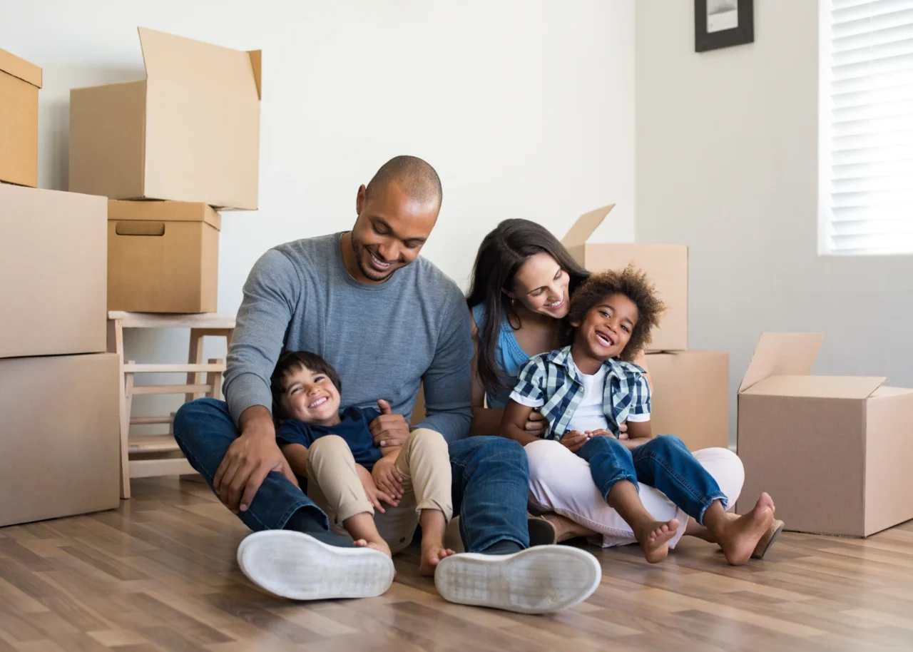 Two parents and two children sit laughing on the wooden floor of an empty new home, surrounded by stacked cardboard moving boxes.