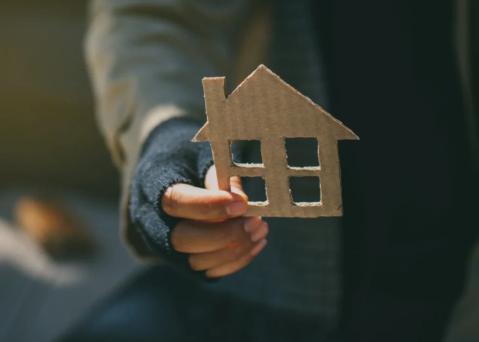 An outstretched hand holds a small cardboard cut-out of a house against a softly blurred outdoor background.