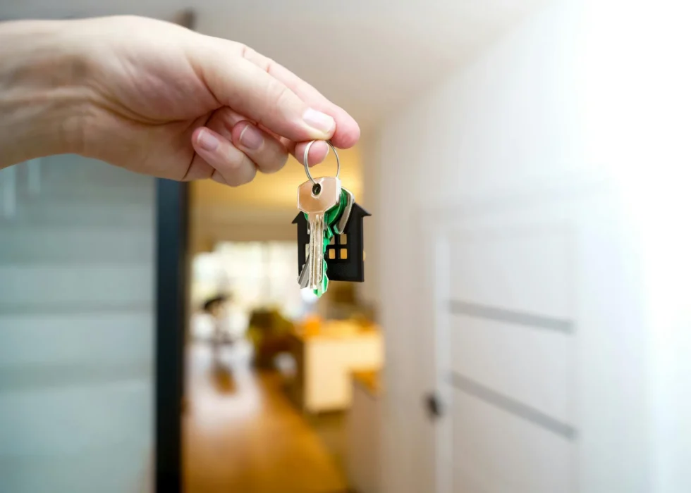 A hand holds a set of keys on a house-shaped keyring, with the softly blurred interior of a home behind them.
