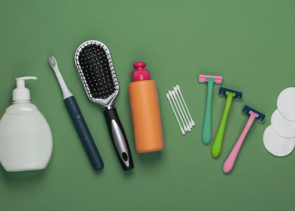 Hygiene supplies laid out in a row on a green background: liquid soap, a toothbrush, a hairbrush, lotion, cotton swabs, disposable razors and cotton pads.