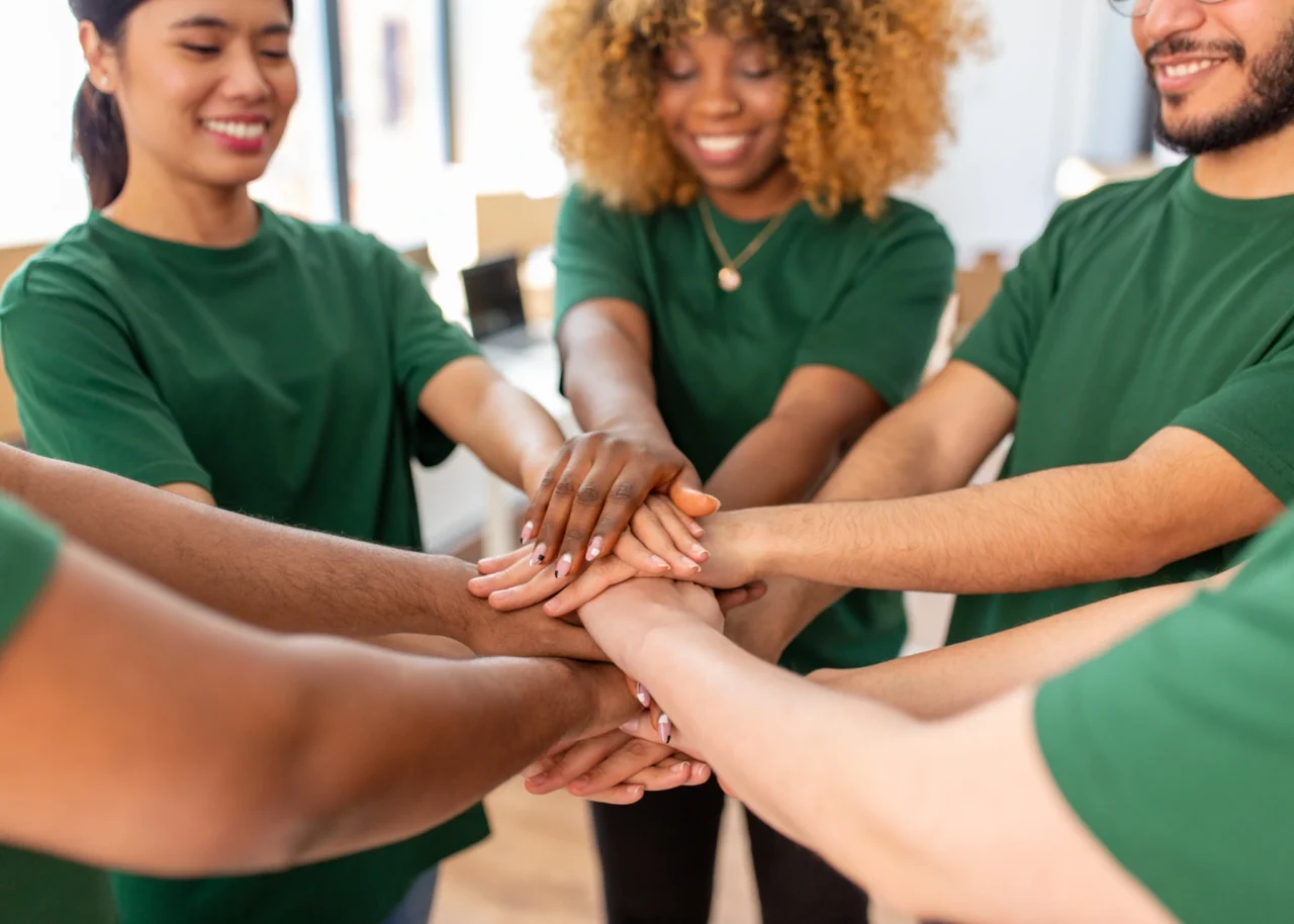 Four volunteers wearing green T-shirts stand in a circle indoors and stack their hands together in the centre, smiling.