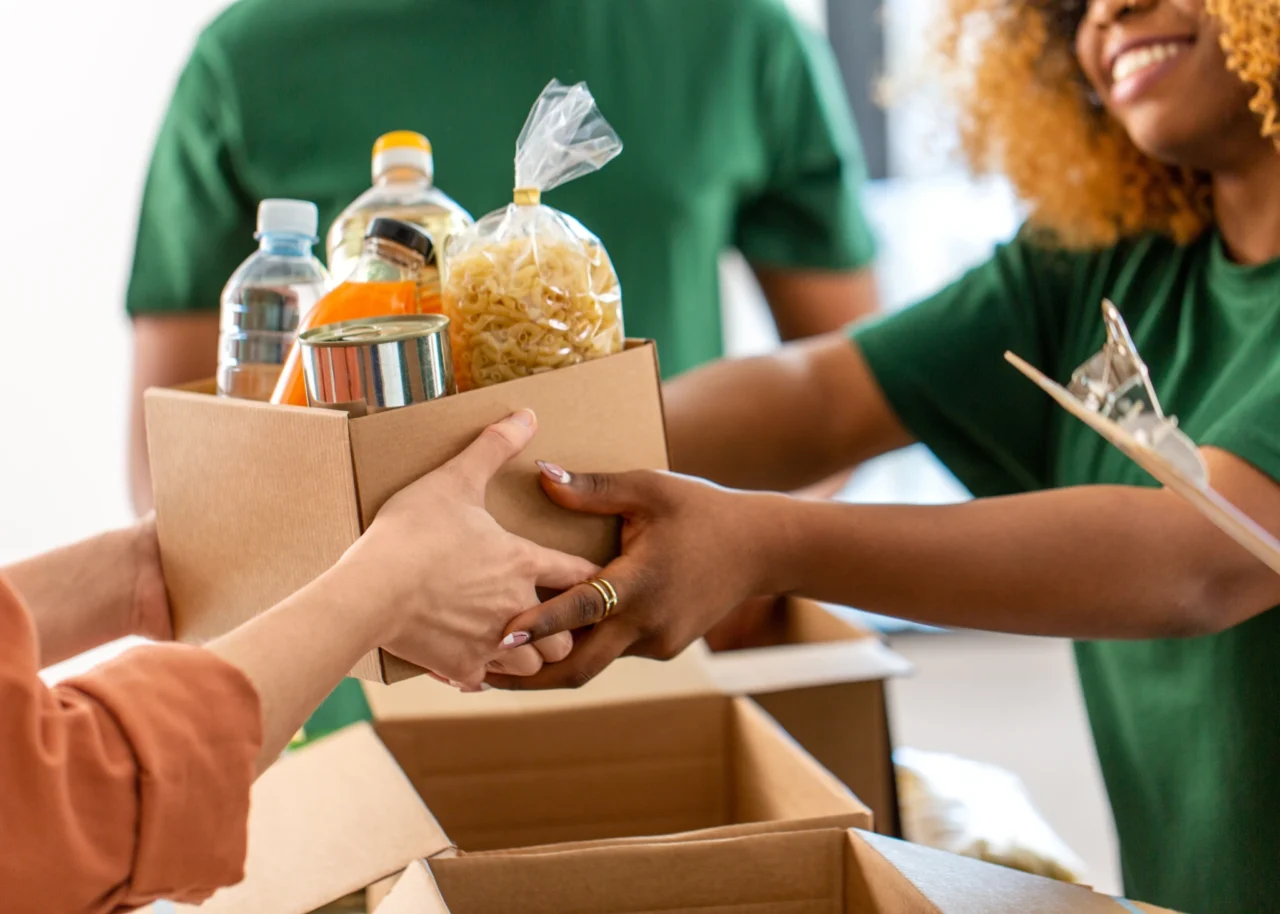 Two pairs of hands pass a cardboard box filled with pasta, canned goods and bottled water between volunteers in green shirts, while another volunteer takes notes on a clipboard.